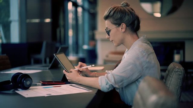 Caucasian woman with electronic pen working with online documents connected to 4g in modern workspace, pensive female freelancer browsing website on digital tablet, e learning concept
