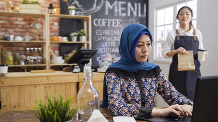 cute korean girl waitress in apron holding disposable coffee cup and paper bag in cafe store walking to muslim woman customer. young elegant islam female client concentrated work on laptop computer