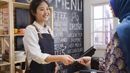 friendly chinese female employee taking payment from muslim customer. young islam lady client giving credit card to pay in cafe bar. smiling waitress hold debit card from arabic guest in coffee shop