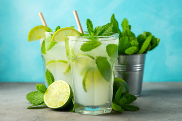 Two homemade lemonade or mojito cocktail with lime, mint and ice cubes in a glass on a light stone...