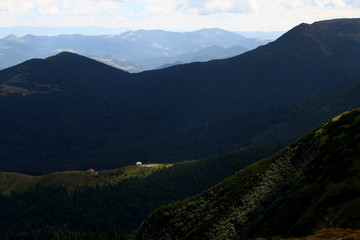 The rays of the sun break through the clouds and illuminate the house standing in a clearing near the edge of the forest among the mountain slopes and peaks. Summer mountain landscape