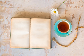 Blank page open book with copy space, tea cup and chamomile flower on wooden desk flat lay background.