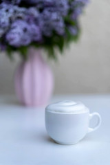 A cup of morning drink with whipped cream on a background of a bouquet of lilacs.