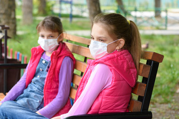 Two girls in medical masks sit on a bench in a park