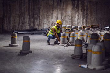 Young bearded worker in vest with helmet on head lifting concrete pillar while crouching in tunnel...