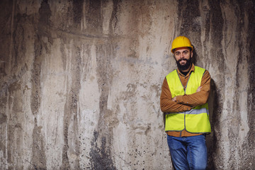 Three quarter length of young smiling attractive bearded worker in vest with helmet on head standing inside of building in construction process with arms crossed, smiling and looking at camera.