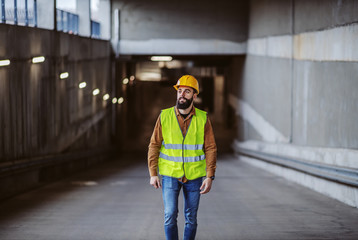 Full length of hardworking attractive bearded worker in vest, with safety helmet on head exiting building in construction precess.