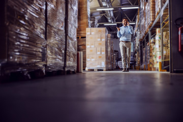 Full length of handsome smiling businessman having phone call with customer while walking in warehouse.