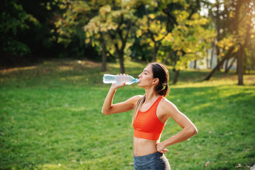 Young gorgeous thirsty slim sportswoman standing with one hand on hip, taking a break after running...