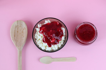Top view of a bowl with cottage cheese and a glass jar with fresh raspberry jam, accompanied by a wooden spoon. Copy space - the concept of proper nutrition, benefits, natural products