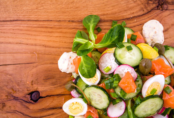 Salad with red fish, fresh vegetables - cucumber and radish, capers, egg, herbs, olives and sauce on a wooden tray, board, dish.