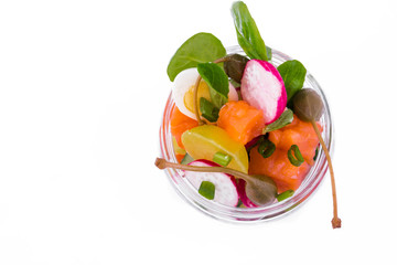 Salad with red fish, fresh vegetables - cucumber and radish, capers, egg, herbs, olives in a glass jar. The object is isolated on a white background.
