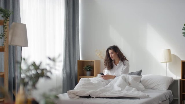 Wide tracking shot of pretty young woman in mens shirt sitting on bed in the morning, listening to music with headphones and smiling while reading something on smartphone