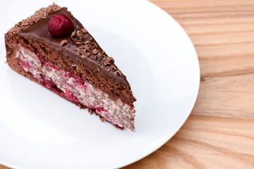 One piece of chocolate cake with vanilla cream and cherry topping served on white plate over light wooden background.