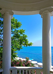 View of the landscape through the columns of a white rotunda on the seashore. Classical arbor with columns in antique style. Seascape with trees, flowers and blue sea.