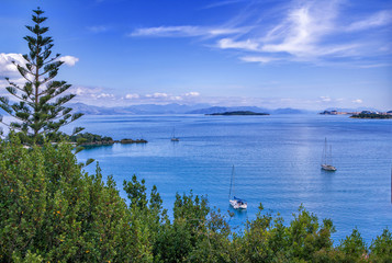 Beautiful summer landscape – calm sea water surface, white yachts, green trees and mountains on the horizon. Corfu Island, Greece.