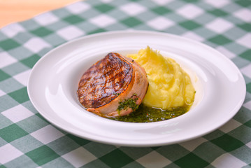 Filet mignon with mashed potatoes and pesto sauce served in a white plate over green plaid tablecloth. Beef steak.