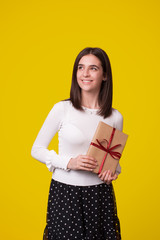Fototapeta premium Young lovely smiling woman is holding a wrapped gift looking far on yellow background.