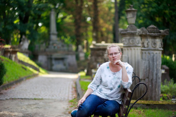 Portrait of a beautiful girl in blue jeans who is sitting on a bench. Lychakiv cemetery. The woman thought. Many graves.