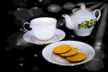 The tea set, consisting of a tea cup with a saucer, a kettle and a plate of biscuits, is located on a black background. side glare of sunlight