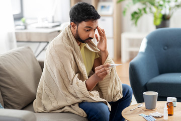 people, health and fever concept - sick indian man in blanket measuring temperature by thermometer...