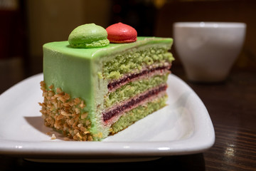a white plate with a piece of green pistachio cake is placed on a wooden lacquered table. close up. white coffee Cup in the background