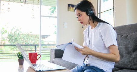 Asian happy woman student wear headphones study online watching webinar podcast on laptop listening learning education course conference calling and write notes look at laptop sit on sofa at home
