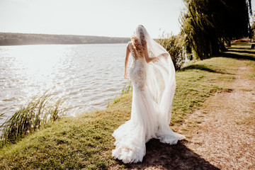 Beautiful bride in a white long lace dress with a long veil and a train of dresses on the grassy shore of the lake