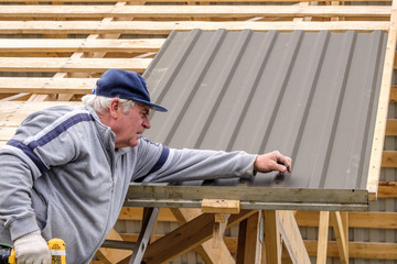 Naklejka na ściany i meble Senior gray-haired Construction man using a screwdriver, fastens a roofing sheet to wooden rafters on the roof of a country house under construction. Physical activity of the seniors.