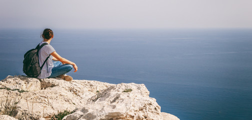 A girl traveler sits on a rock and admires the blue boundless sea, freedom, travel, unity with nature, banner panorama format