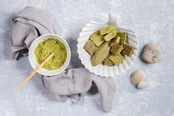 Bowl of Matcha powder, plate as shell with treats containing matcha, napkin and stones on light grey background.