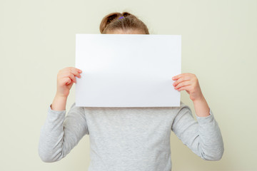 Clean sheet of a paper in hands of child covering her face on yellow background. Mockup sheet of paper in child's hands.