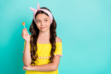 Portrait of minded creative sly child girl hold lollipop look copyspace think thoughts plan weekends wear yellow stylish trendy dress isolated over teal color background