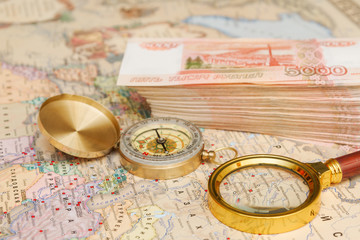 A compass with a bundle of big money and a magnifying glass are on the map