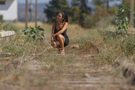 Mujer morena, modelo, con vestido de negro y chancletas, con gafas de sol agachada en un paisaje verde y hierba seca 