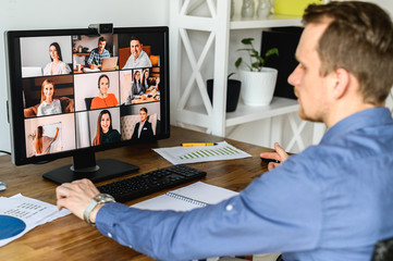 Virtual conference with employees. A young man in formal shirt using pc for video call, he has video meeting with several people together. Back view. Remote work concept.