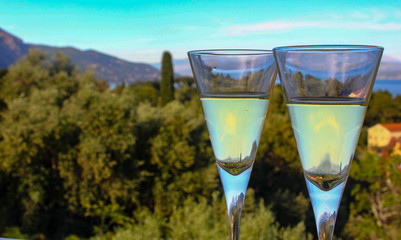 Two Glasses of white wine on seascape background with sun sky mountains and cypress tree landscape reflected in alcohol drink.  An evening cocktail before romantic dinner outdoors on holiday balcony.