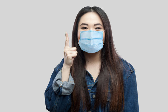Idea! Portrait of exicted beautiful brunette asian girl with surgical medical mask in blue denim jacket standing and looking at camera , finger up. indoor studio shot, isolated on grey background.