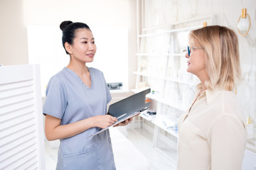 Friendly Asian cosmetologist with clipboard providing client with information about procedure in beauty salon