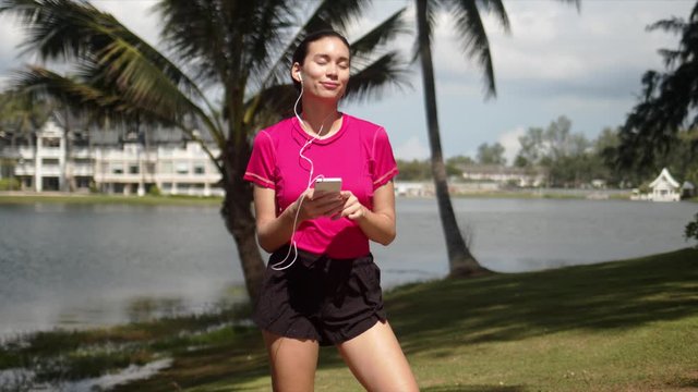 Young cheerful sportive female wearing shirt and shorts using smartphone and earphones