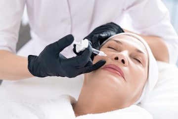 Close-up of unrecognizable cosmetologist using syringe while injecting filler in women's face to rejuvenate her skin