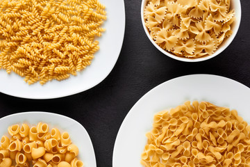 Variety of raw pasta in white plates and bowls on brown textured background