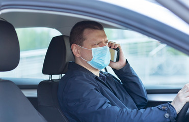 A young man in a protective mask is sitting in a left-hand drive car and  calls on his mobile phone, looking at the camera. Driver in a medical mask. Coronavirus covid-19, pandemic. Virus protection 