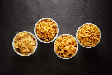 Variety of raw pasta in white bowls on brown textured background