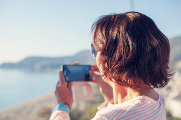 Attractive retired woman 50-60 years old travels and takes photos on a smartphone at the viewpoint...