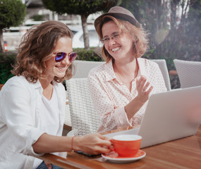 Two happy young beautiful women girls girlfriends sitting in cafe drink coffee with laptop on table