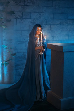 young woman queen stands in dark night room mysterious castle. Lady elf princess holding candlestick in hands, candles burn. Blue silk medieval dress, elegant clothes vintage cloak hood, silver tiara