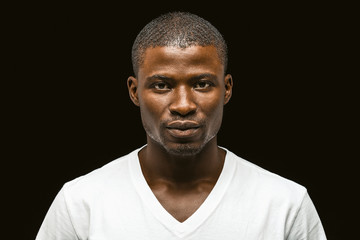 Serious African american man looking at the camera, attractive dark-skinned young guy in a white t-shirt posing expressing confidence isolated on black background. Toned image