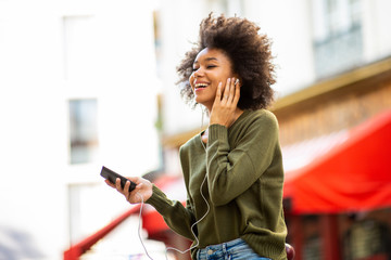 Side of happy young black woman listening to music with earphones and smart phone in city