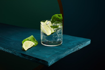 old fashioned glass with fresh drink, mint leaf and lime slice on blue wooden surface on green and...